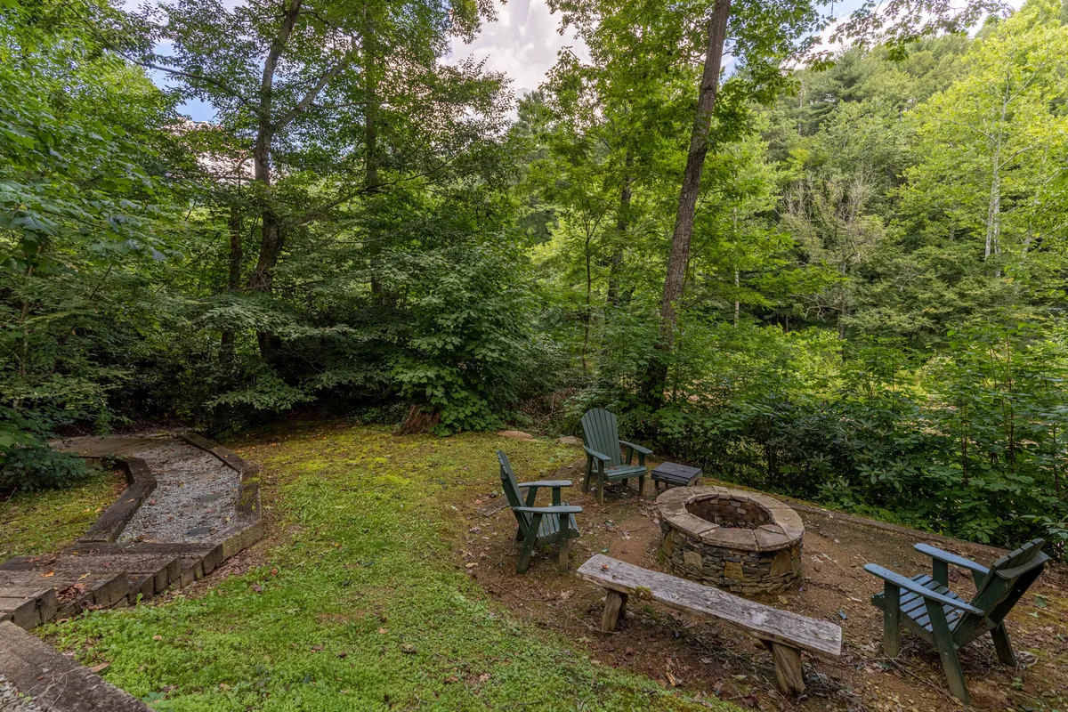 This inviting scene features a stone fire pit encircled by Adirondack chairs, set along a stone path amid vibrant green landscaping. The surrounding trees and open space suggest a welcoming area for evening gatherings and storytelling under the stars.