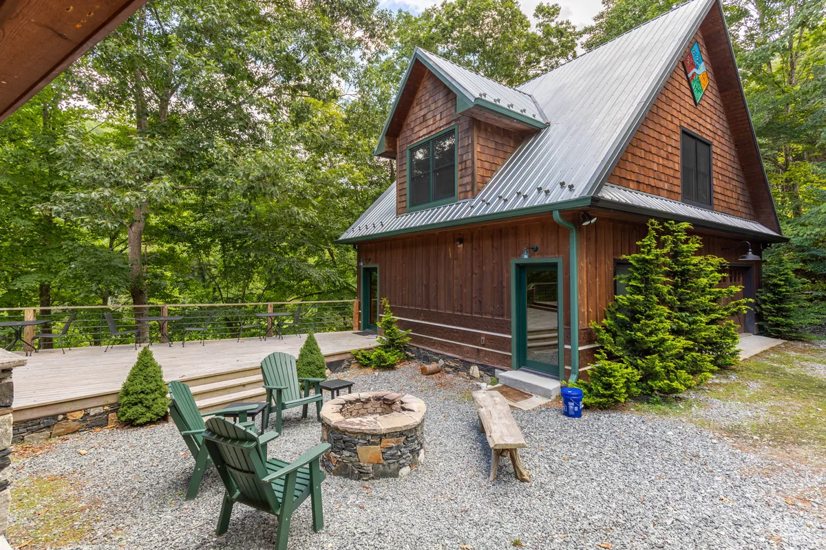 A cozy outdoor area featuring a fire pit surrounded by Adirondack chairs on a wooden deck, nestled in lush greenery. The inviting setup overlooks the landscape, perfect for evening gatherings, stargazing, or enjoying the sounds of the river nearby.