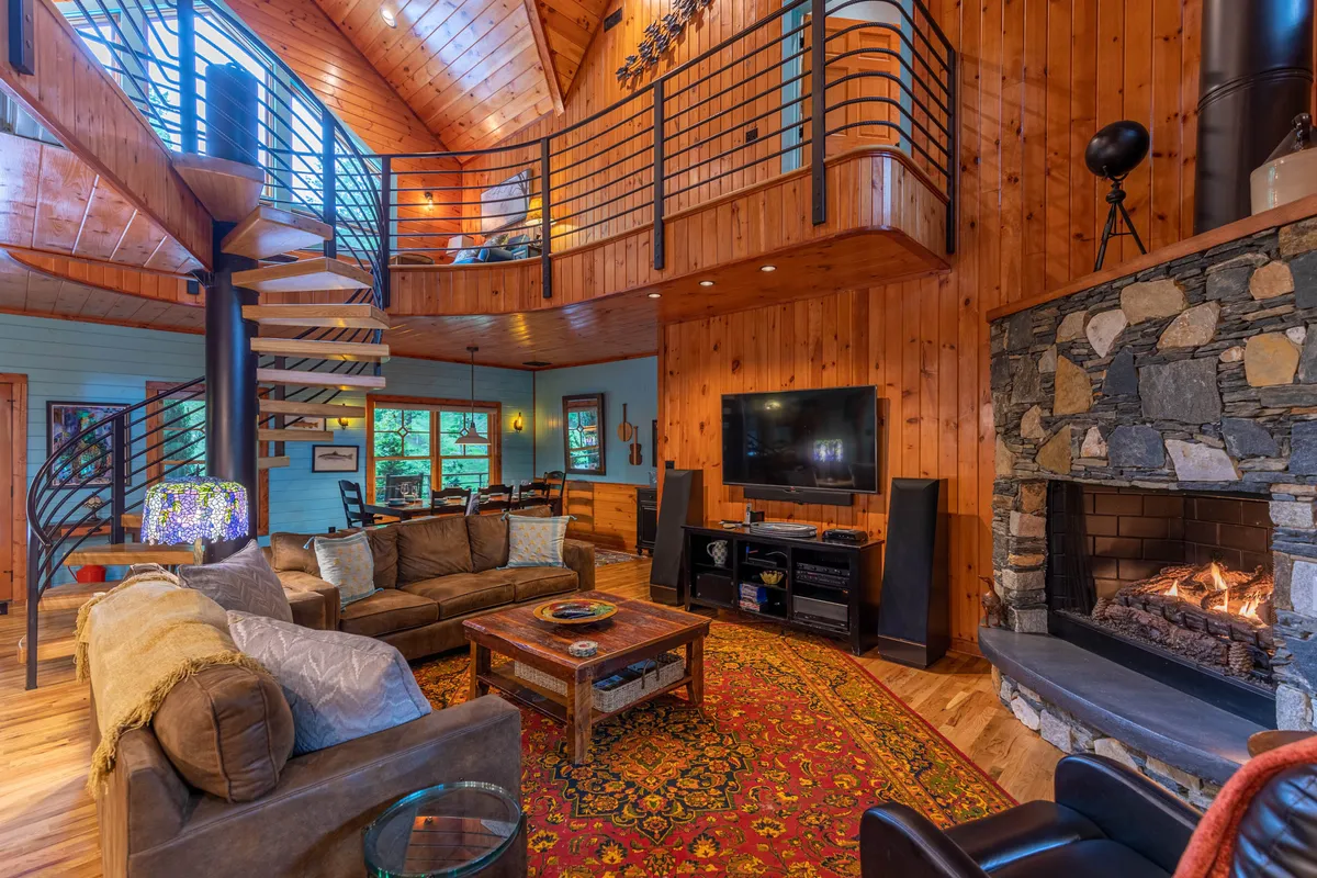 A cozy living room with a spiral staircase, plush sofa seating arranged around a fireplace and a large TV, all accented by a stylish rug and warm wood finishes. Large windows offer glimpses of the outdoors, emphasizing comfort and togetherness.