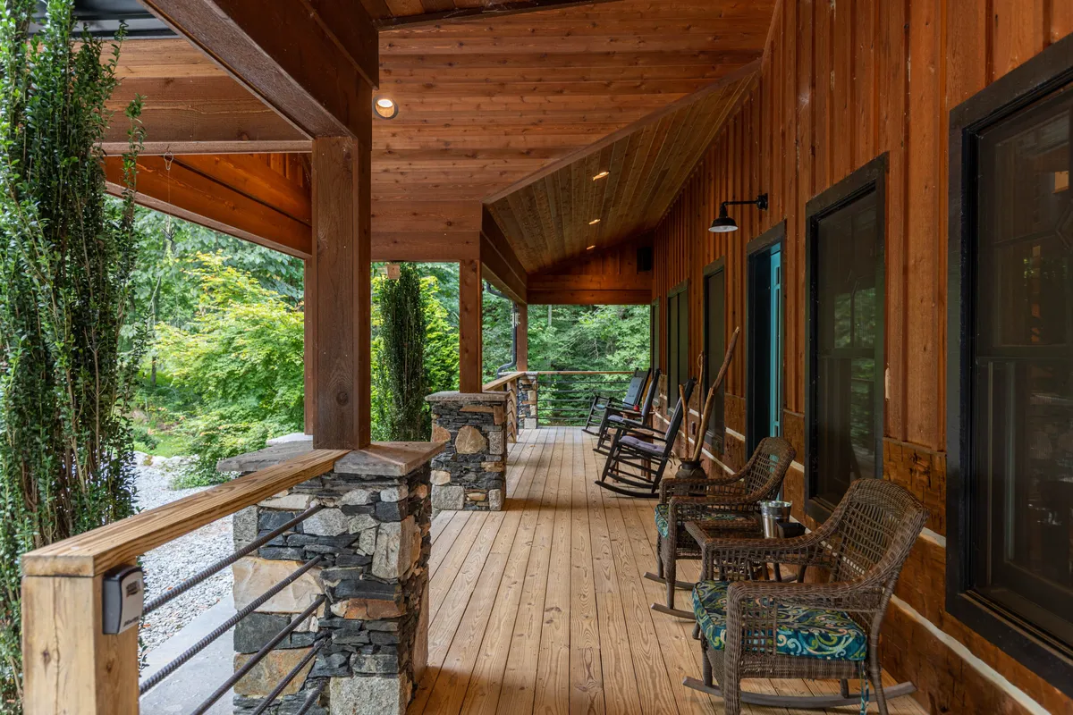 A peaceful exterior porch scene with wooden rocking chairs, stone railing, lush greenery, and exposed wooden beams. The inviting chairs face outward, ideal for soaking in sunset views or quietly admiring the surrounding landscape.