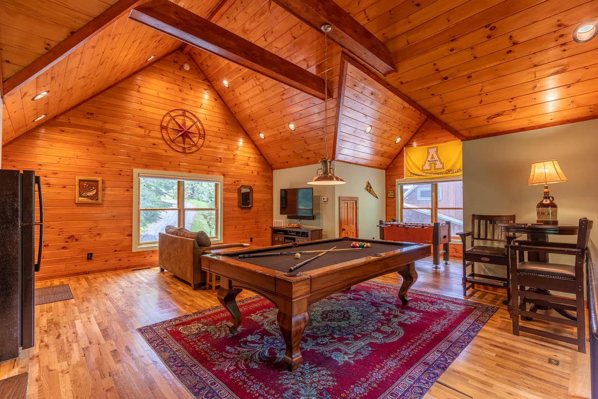 This image features a vibrant game room with a pool table as the focal point, wooden beams overhead, a cozy rug, comfortable seating, and decorative items that contribute to a relaxed and playful atmosphere. The space looks inviting for socializing, playing games, and spending time indoors.