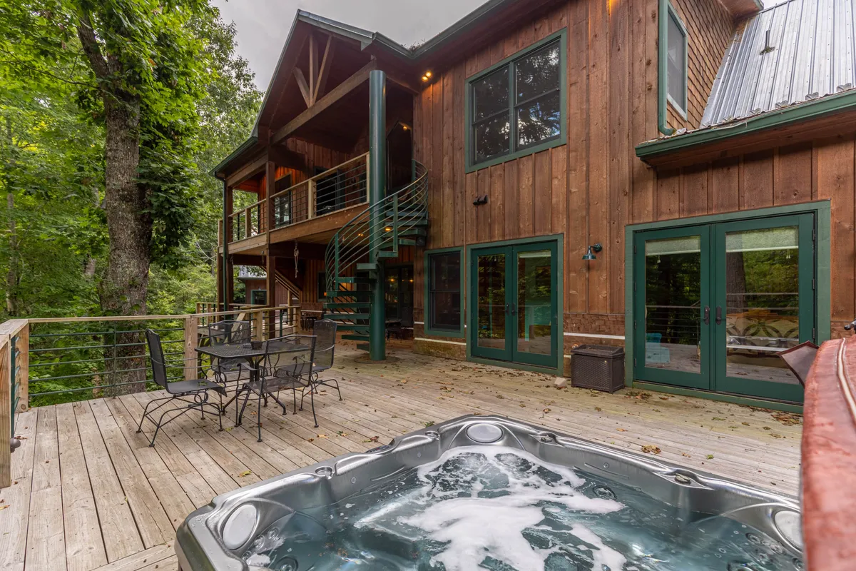 The image showcases a spacious wooden deck overlooking the surrounding forest, featuring a built-in hot tub, an outdoor dining table, and a striking spiral staircase that leads to an upper level. The deck is bordered by railings and surrounded by lush greenery, evoking a sense of seclusion and connection with nature.