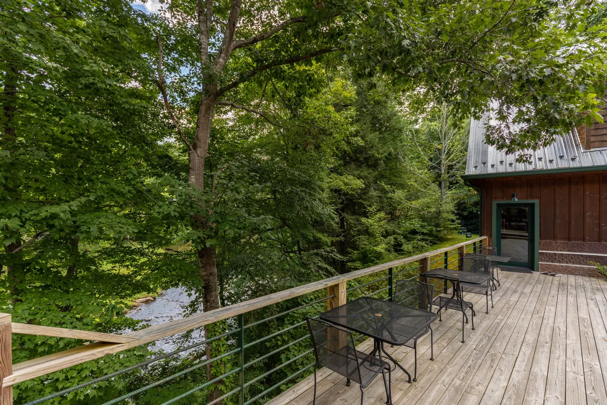 A wooden deck perched above the river, with tables and chairs for outdoor relaxation. The scene highlights the river flowing beside the property, framed by trees and bathed in peaceful natural light.