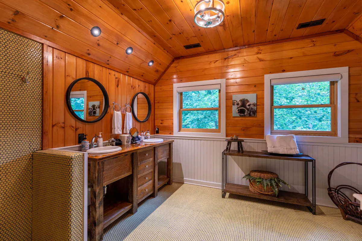 A double wooden vanity with dual mirrors, large windows letting in sunlight, and a decorative basket in a warm, rustic bathroom setting. The space feels generous and perfect for morning routines or evening relaxation.