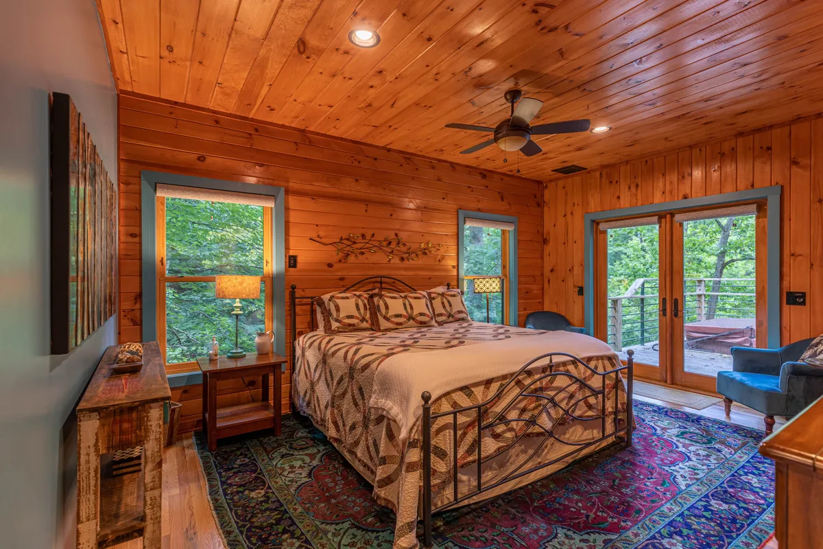 This image features a king bed with a decorative quilt, a wooden ceiling, expansive windows with tranquil nature views, an accent chair, and a side table with a lamp. The setting is spacious, serene, and highlights comfort and privacy, echoing the atmosphere of a personal sanctuary.