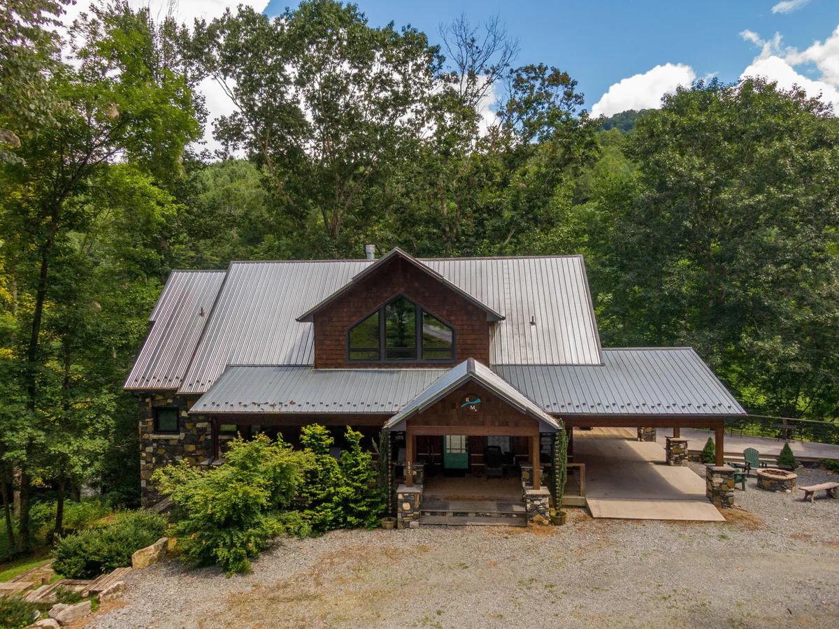This image showcases the cabin's striking wooden facade and metal roof, set against lush greenery. The expansive front porch invites relaxation, with cozy seating areas perfect for taking in sunset views. The surrounding landscape includes hints of a fire pit area and dense woods, promising tranquil moments and nature immersion.