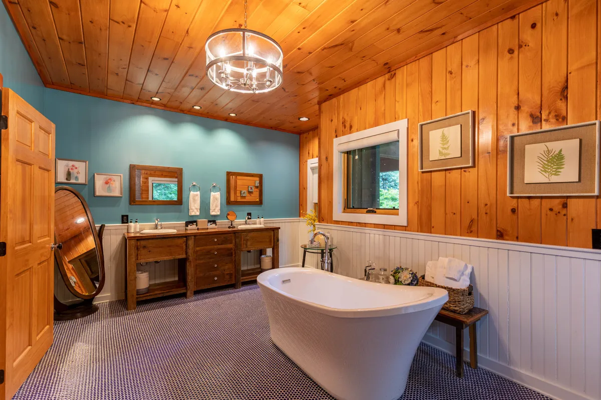 A spa-inspired ensuite bath featuring a freestanding soaking tub, wooden vanity, stylish chandelier, and framed artwork. The bath area is filled with natural light and exudes a calm, rejuvenating vibe.