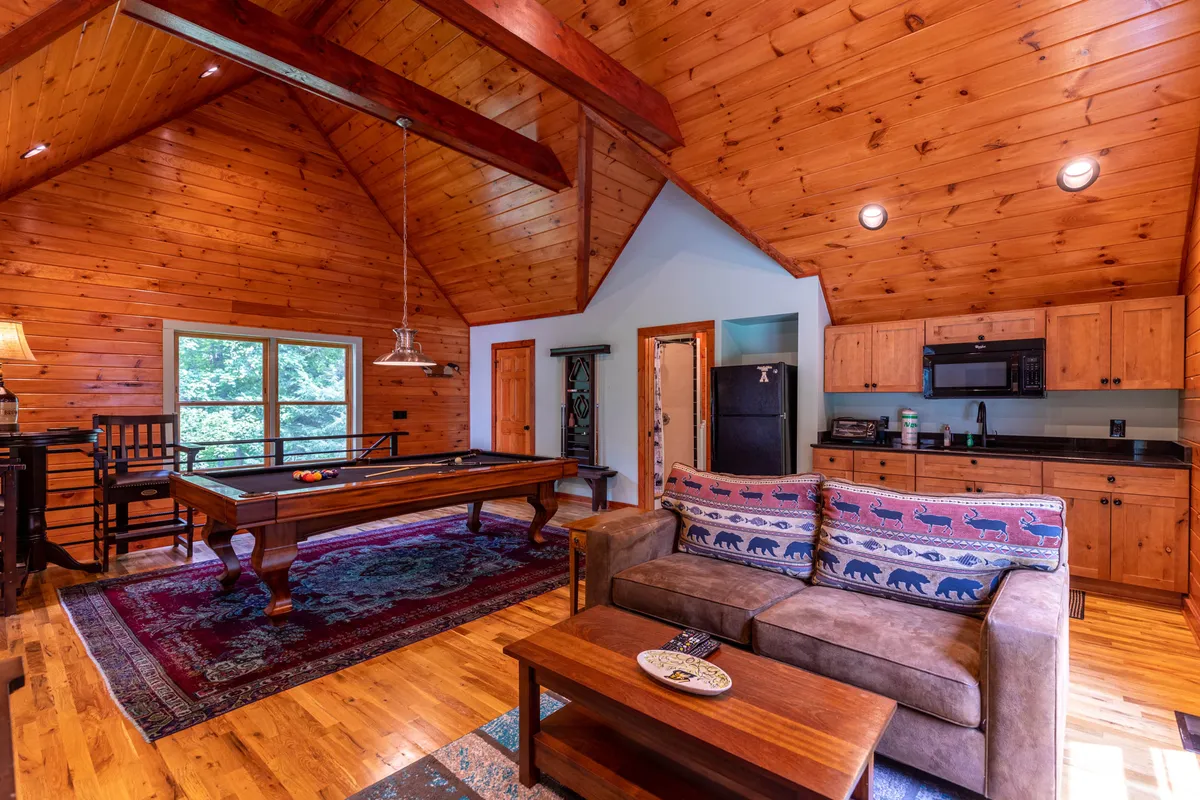 Inside, a spacious game room includes a pool table, sofa, wooden ceiling, and large windows letting in natural light. The comfortable setup encourages friendly competition, family game nights, and relaxed socializing.