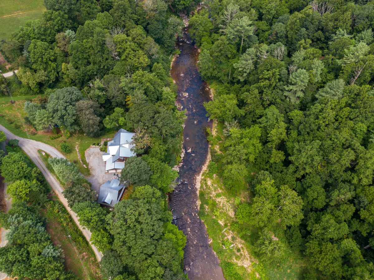 This image captures the tranquil Watauga River flowing beside the house, hugged by dense trees. The house is visible in the background, emphasizing the close proximity to the river and the surrounding natural landscape. The scene is serene, suggesting both opportunities for fishing and moments of peaceful relaxation after outdoor activities.
