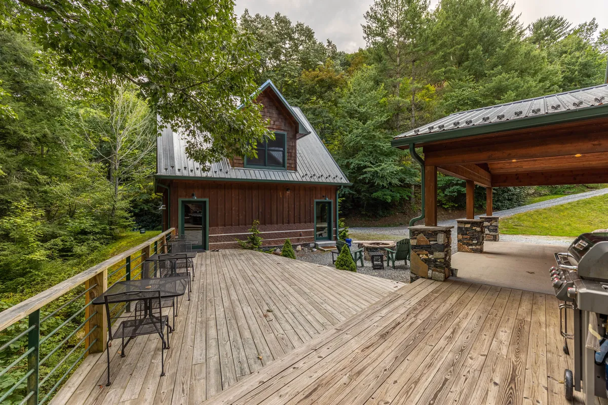 A wooden deck attached to a two-story cabin offers an outdoor seating area and a stone barbecue, nestled amongst trees. The scene is set for barbecuing, relaxing in nature, and enjoying the natural surroundings with family, friends, and pets.