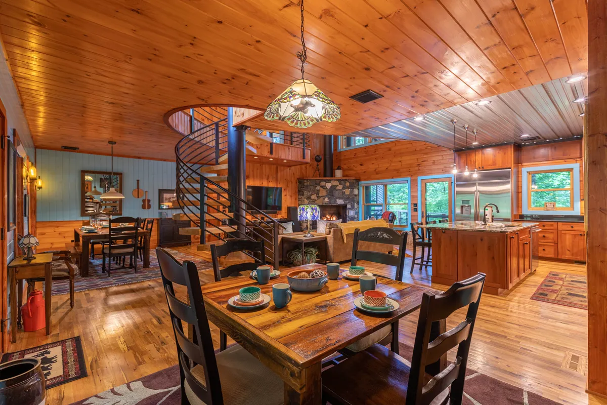 A spacious, open-plan great room featuring a central dining table, a dramatic spiral staircase, a locally-sourced stone fireplace, and a large kitchen island. The area is warmly lit and decorated in mountain-modern style, with hardwood floors and expansive windows letting in natural light.
