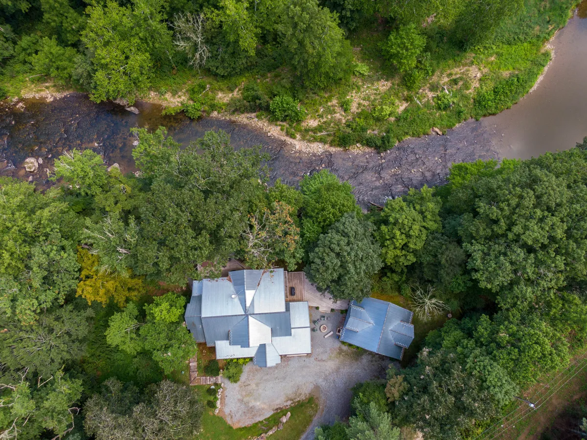 This image features a peaceful, tree-lined riverbank with the house set just steps from the water. The outdoor seating area is visible, offering a direct connection to the river. The atmosphere is tranquil, ideal for fishing, relaxing by the campfire, or enjoying the hammock under mature trees.