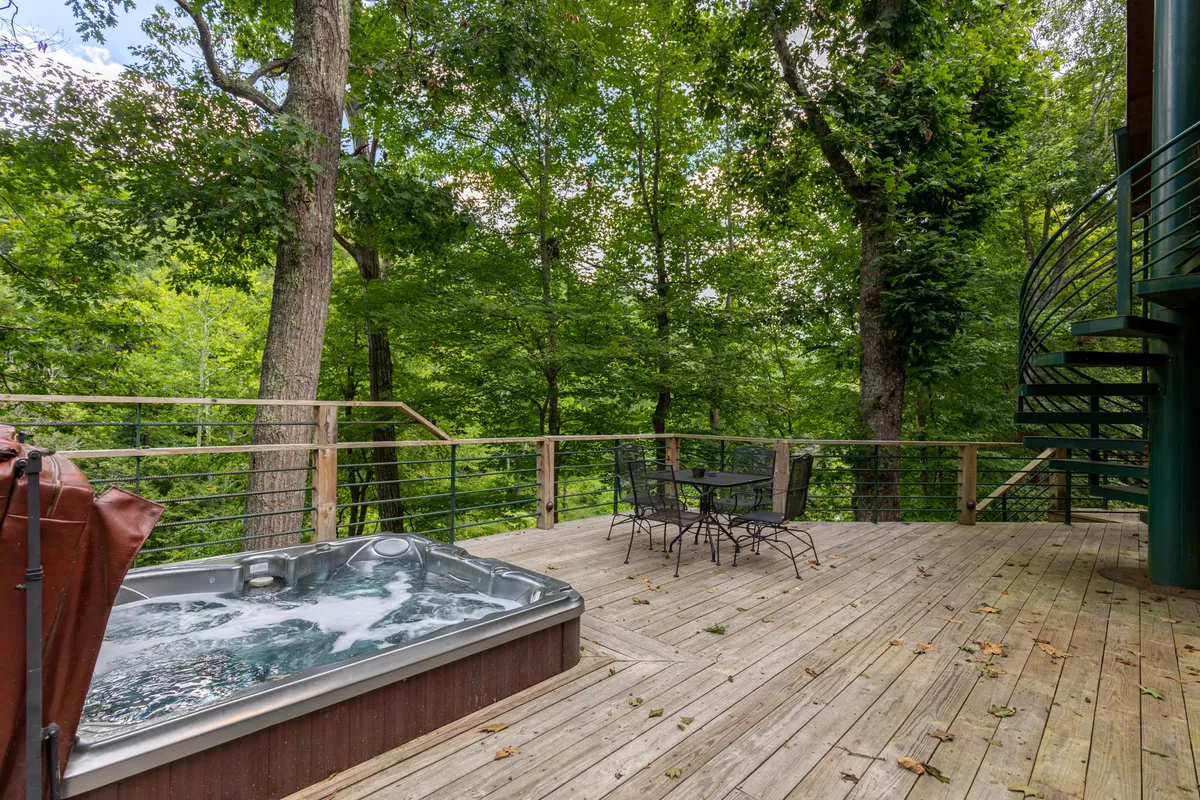 A tranquil deck setting with a bubbling hot tub, an outdoor dining set, and mature trees providing a natural backdrop. The space is designed for both relaxation and socializing, offering room for families, friends, and furry companions to unwind together.